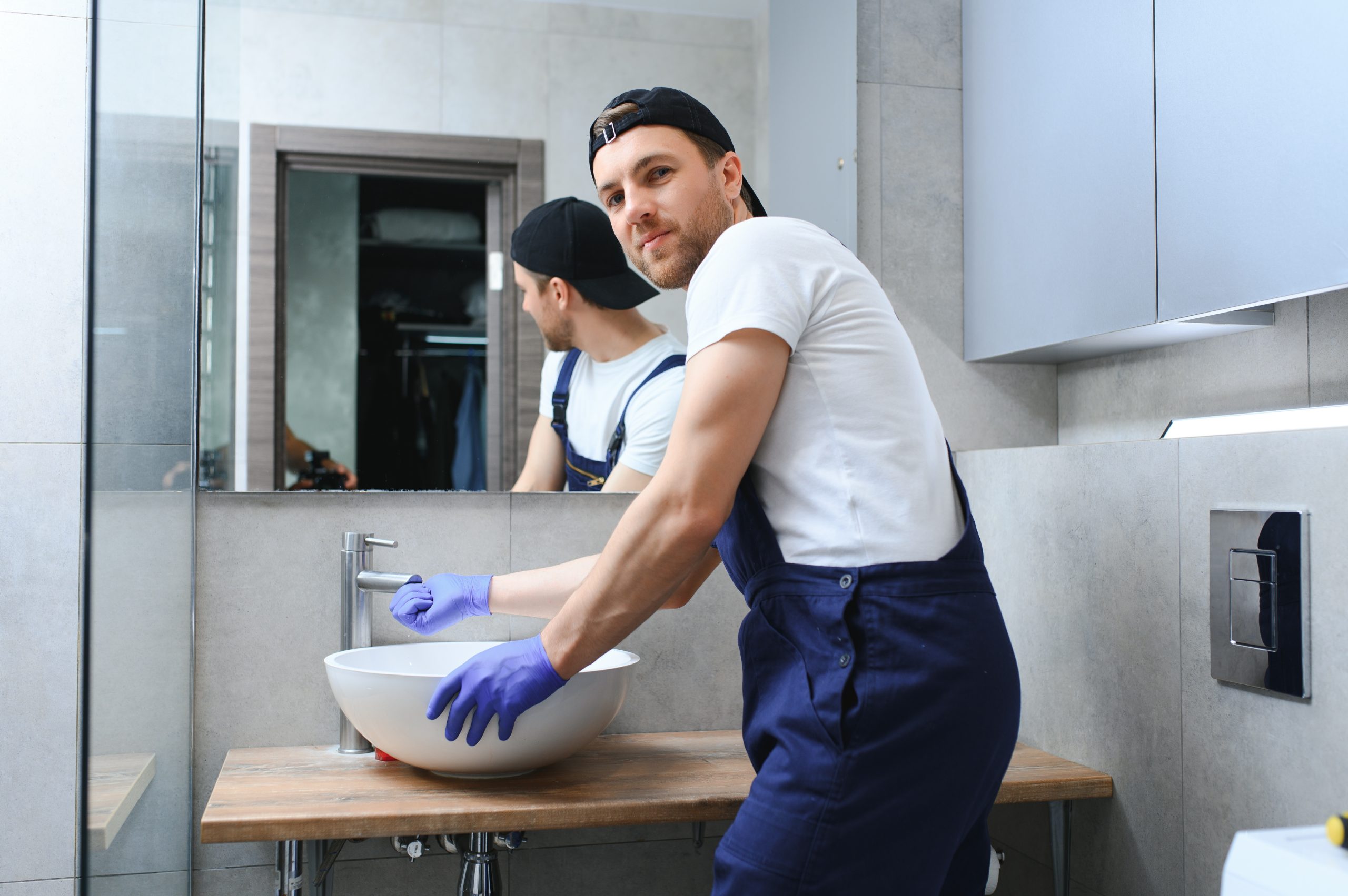 professional plumber, male worker in uniform installing sink and water pipe in new apartment.