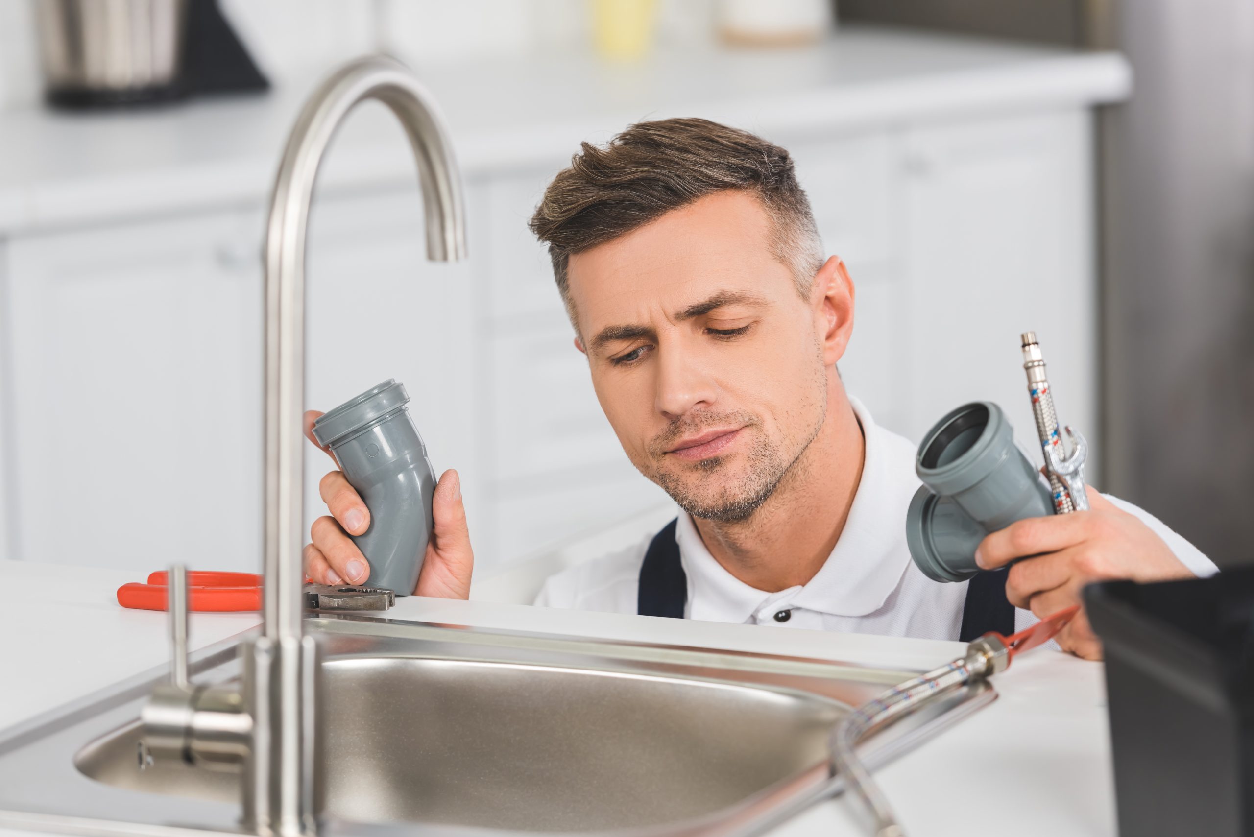 thoughtful adult repairman holding pipes and tools while repairing faucet at kitchen