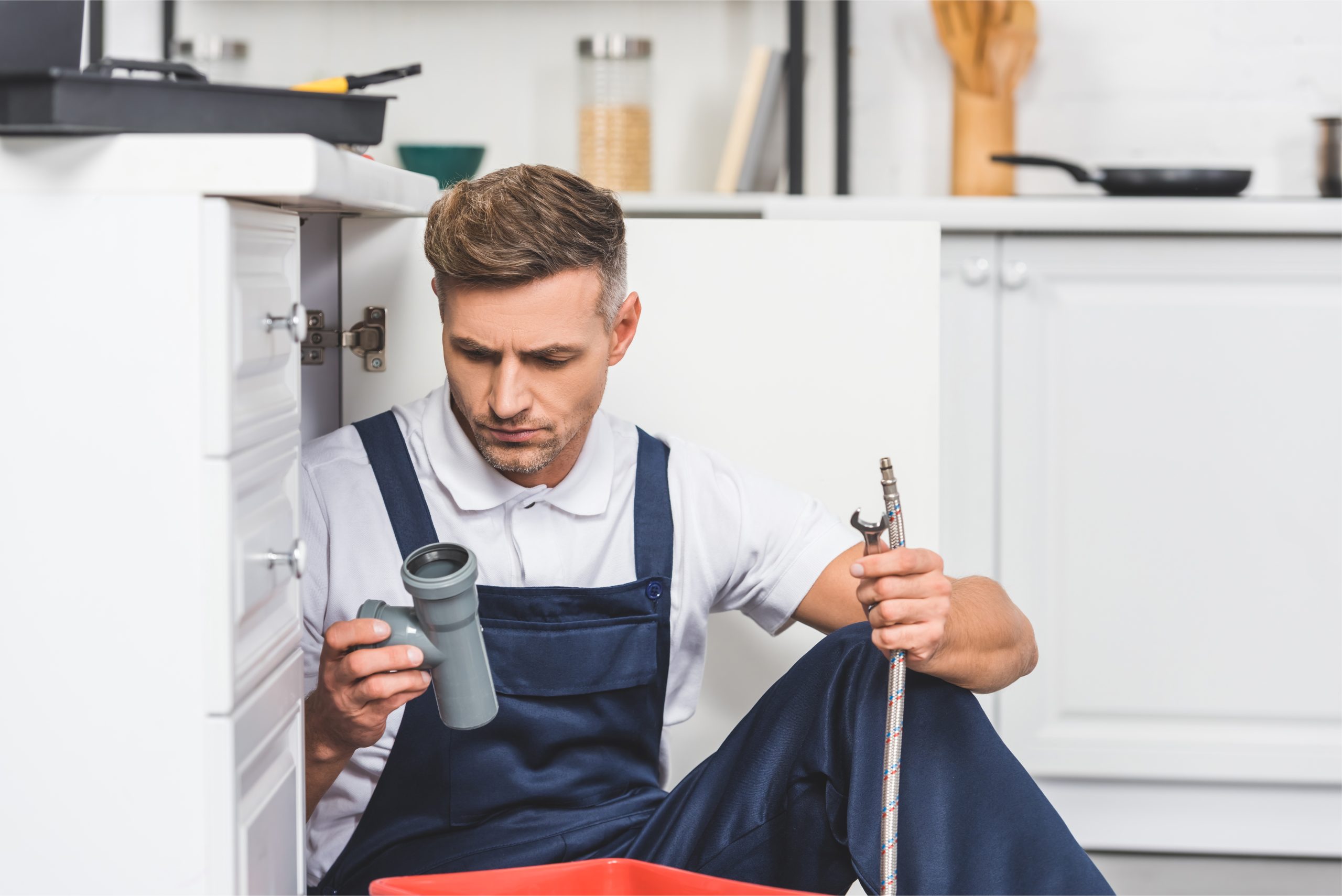 thoughtful adult repairman sitting under sink and holding pipes for repairing at kitchen